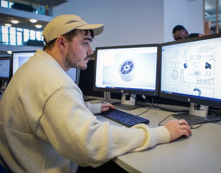 A student wearing a white cap working on a dual-monitor CAD setup, designing a mechanical component on one screen and a technical drawing on the other. A student wearing a white cap working on a dual-monitor CAD setup, designing a mechanical component on one screen and a technical drawing on the other.